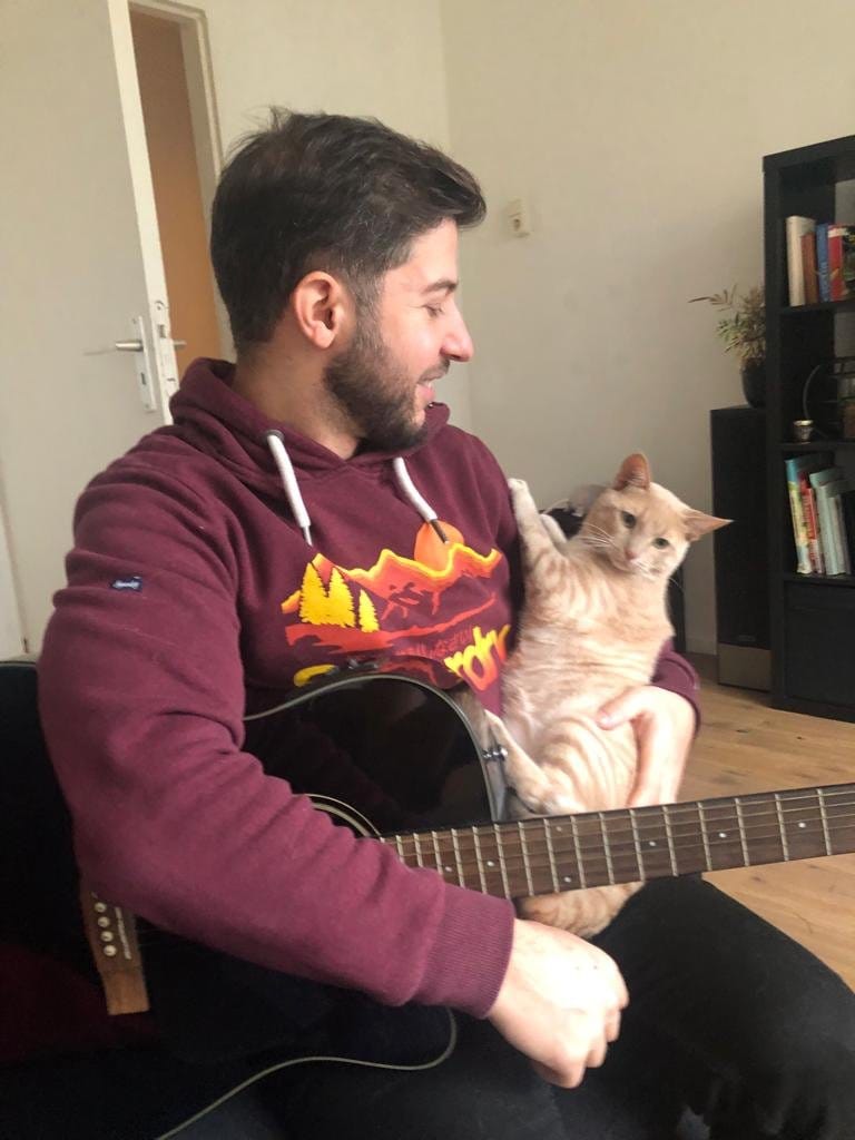 A man in a maroon hoodie plays guitar with a cream and ginger tabby sitting beside him.