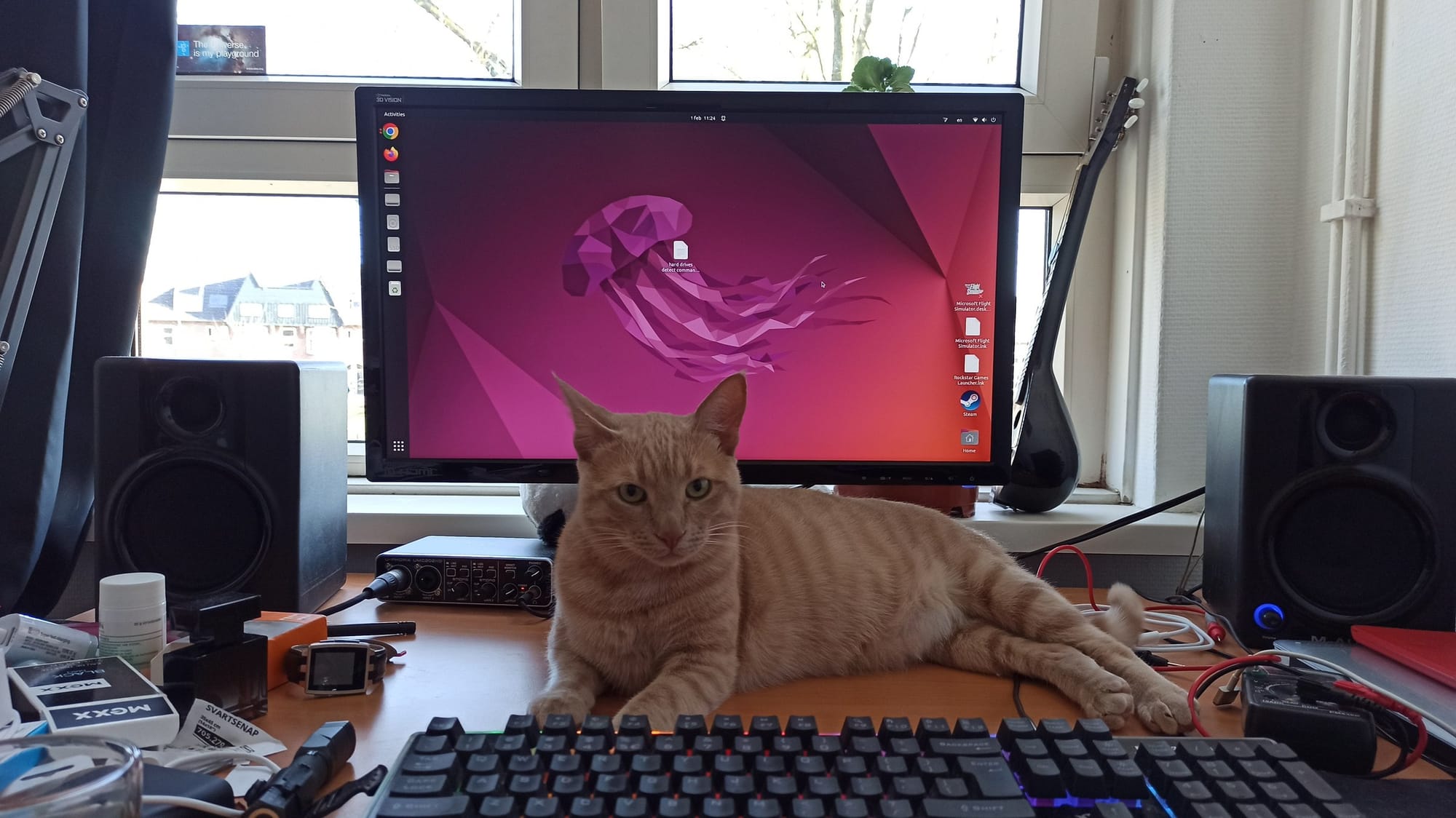 A ginger cat lies stretched out on a keyboard in front of a computer monitor displaying a colourful desktop background.