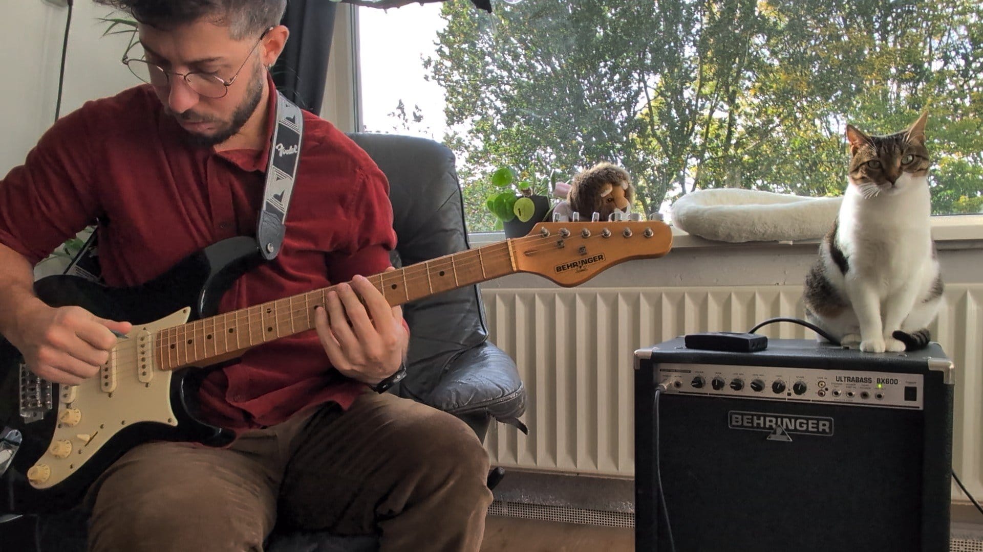 A man plays a guitar next to an amplifier while a cream and ginger tabby sits attentively nearby.