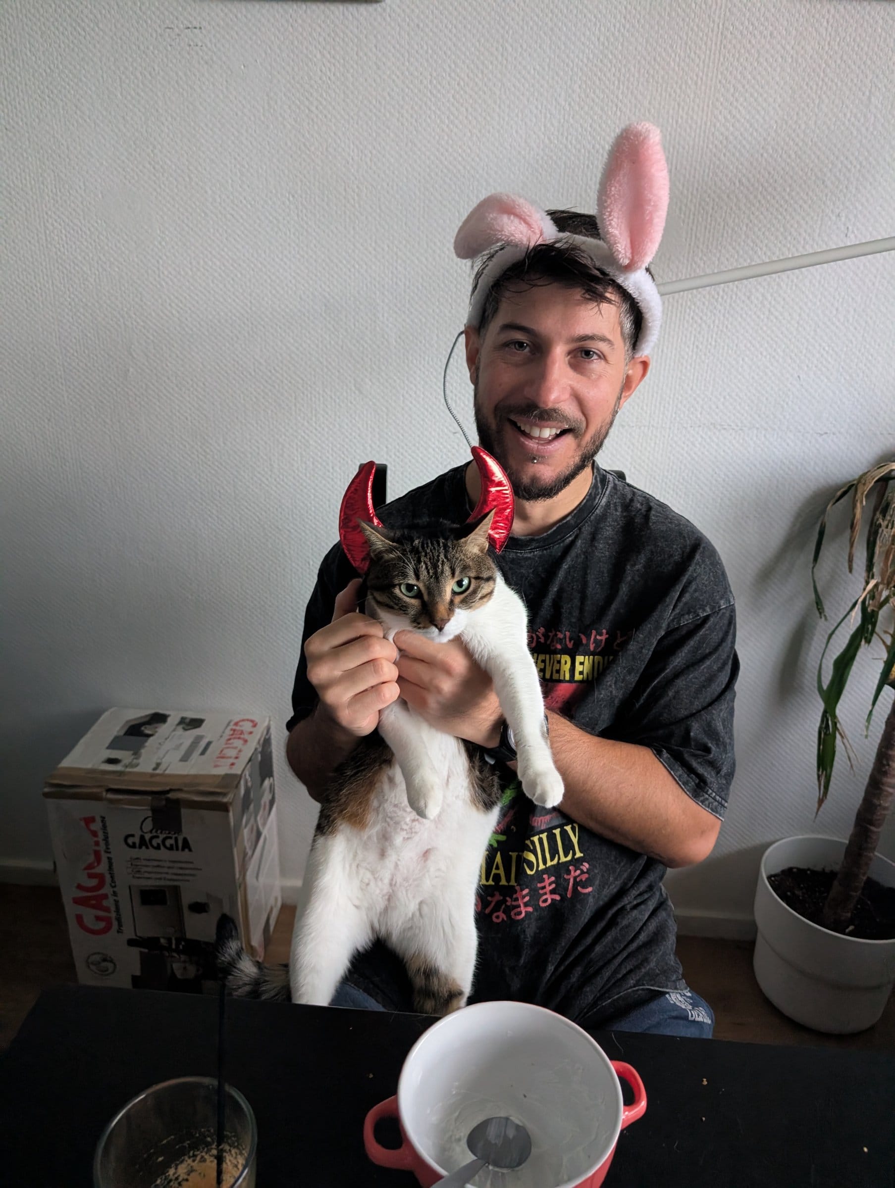 A smiling man wearing bunny ears holds a brown and white tabby wearing devil horns.