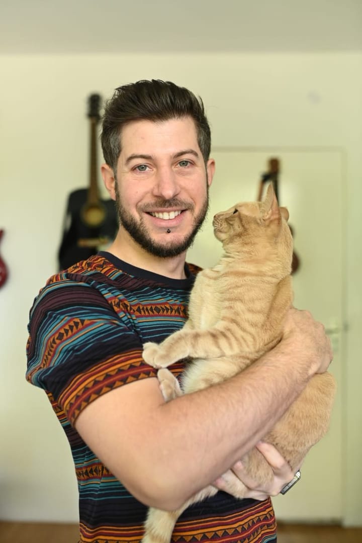 A smiling man holds a ginger cat in his arms indoors, with guitars visible in the background.